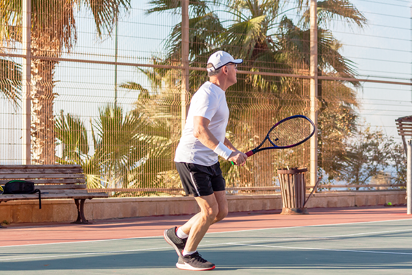 An elderly man plays tennis on an outdoor court 